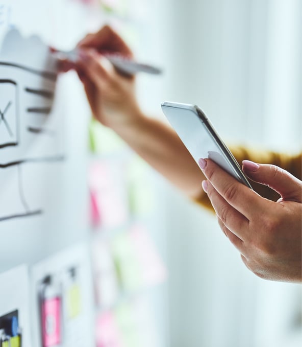 Hände einer Person, die mit Links ein Smartphone hält und rechts auf Whiteboard schreibt 