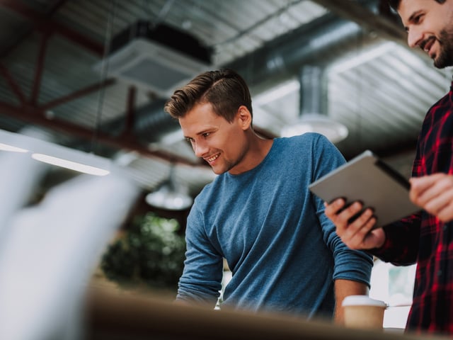 Zwei lächelnde Arbeitskollegen in Lagerhalle, die auf Computer- und Tablet-Bildschirme schauen 