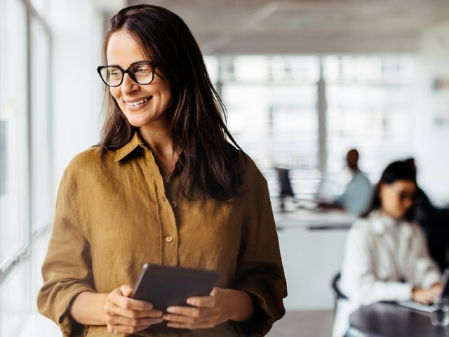 Woman in glasses looking out the window smiling with tablet in her hands