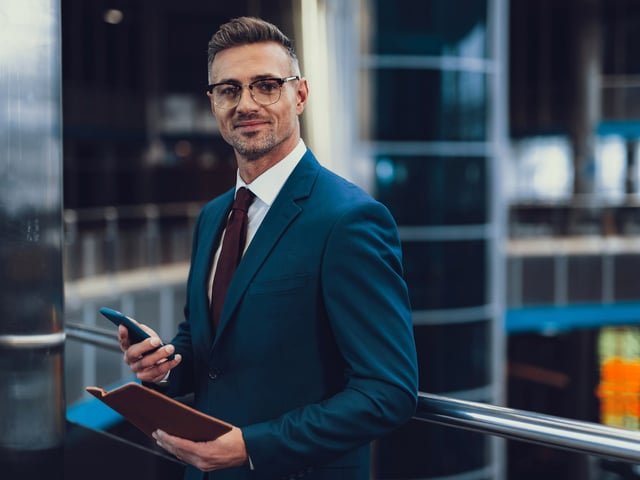 Man in business suit smiling and holding book in left hand and smartphone in right hand 