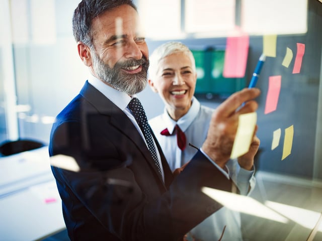 Weibliche und männliche Arbeitskollegen, die lächelnd auf ein Post-it an einer Tafel zeigen 