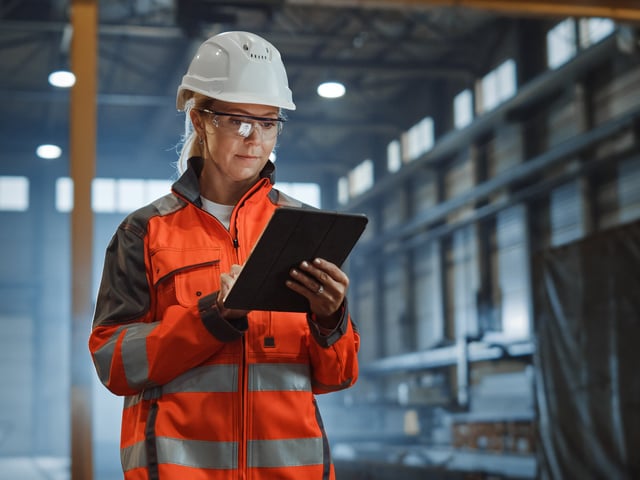 Frau mit Schutzanzug und Helm, schaut auf ein Tablet in ihren Händen 