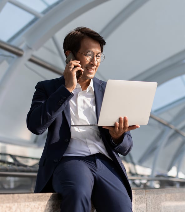 Man in business suit leaning against wall holding laptop and talking on smartphone 