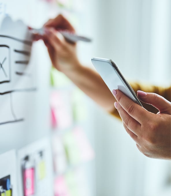 Hände einer Person, die mit Links ein Smartphone hält und rechts auf Whiteboard schreibt 