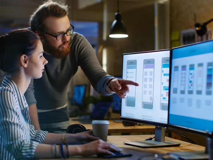 Male working colleague pointing at computer screen of female working colleague sitting at desk 
