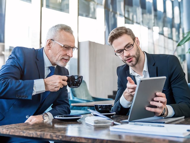 Male working colleague showing another working colleague wo is drinking coffee something on his tablet 