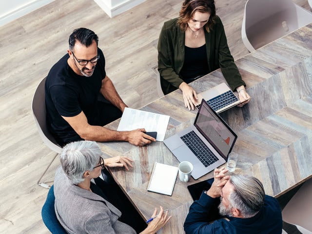 Four work colleagues sit around a meeting table and discuss, laptops and notepads lie on the table 