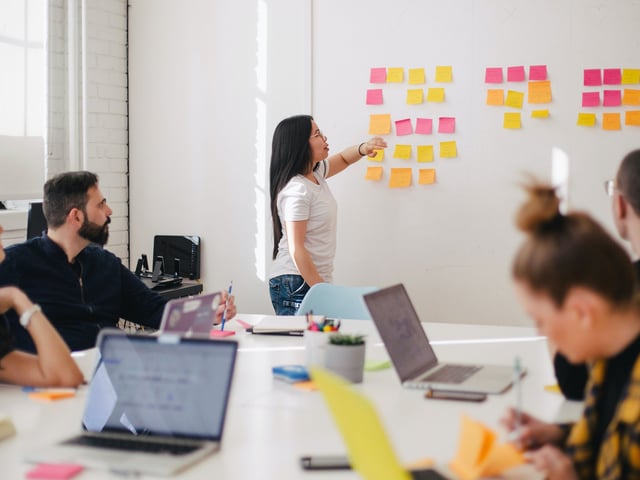 Female working colleague shows sticky-notes on whiteboard while several colleagues sit around a table with laptops in front of them 