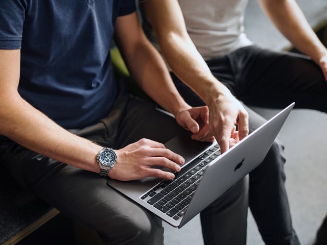 Zwei Arbeitskollegen sitzen nebeneinander, der eine hält einen Laptop auf dem Schoss, der andere deutet auf etwas auf dem Display 