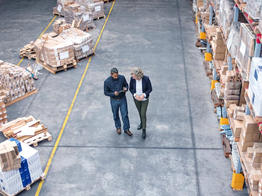 Two men with helmets walking through storehouse talking 