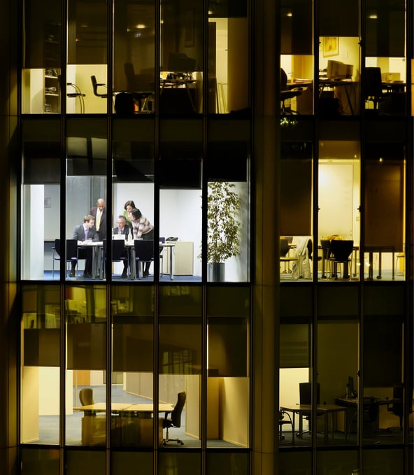 Drei Stockwerke eines Gebäudes in der Nacht, im beleuchteten mittleren Stockwerk sitzen zwei Personen an Schreibtisch 