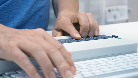 Man touching Braille keyboard with index finger of left hand while typing on standard keyboard with right hand 