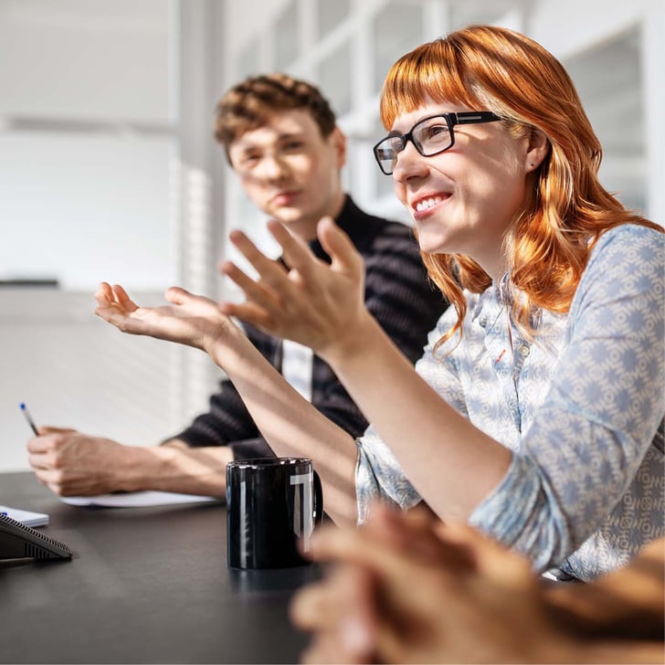 Woman at business meeting smiling and holding her hands up while male colleague observes her from the side 