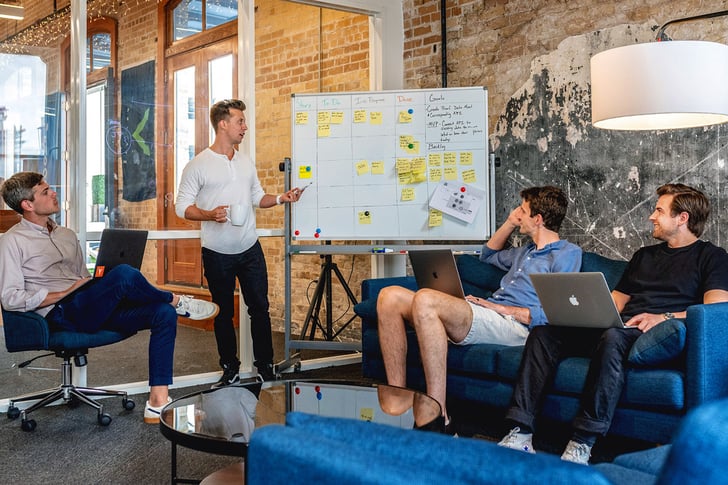 working colleague shows sticky-notes on whiteboard while several colleagues sit around a table with laptops in front of them