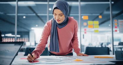 A woman in a hijab working on a project at a desk in a modern office, reviewing documents and writing notes with colorful sticky notes in the background.