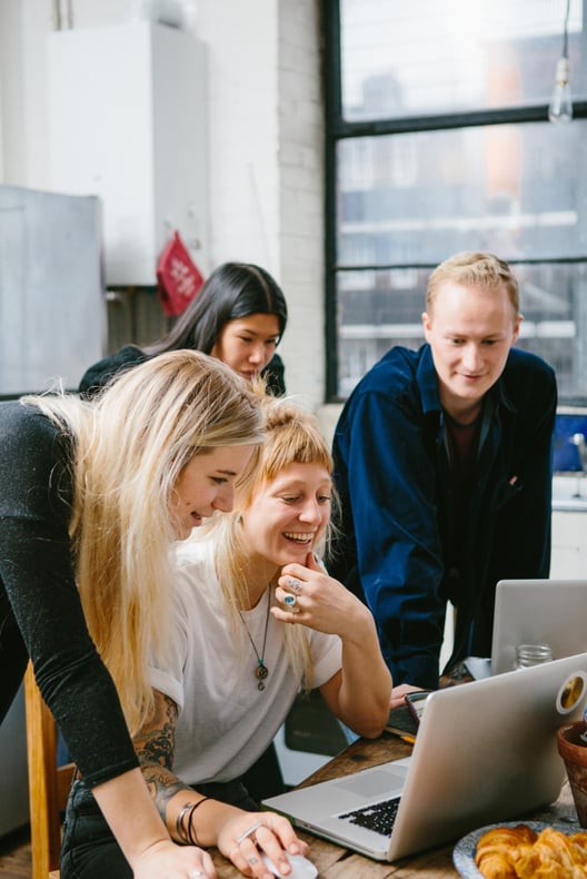 Drei weibliche und ein männlicher Arbeitskollege schauen lächelnd auf einen Laptop-Bildschirm 