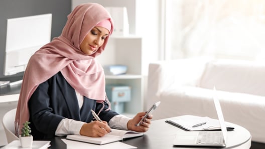 Woman sitting in front of laptop, holding smartphone in left hand and taking notes with right hand   