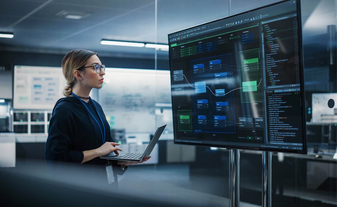 young_woman_holding_a_laptop_computer_and_looking_at_a_big_screen