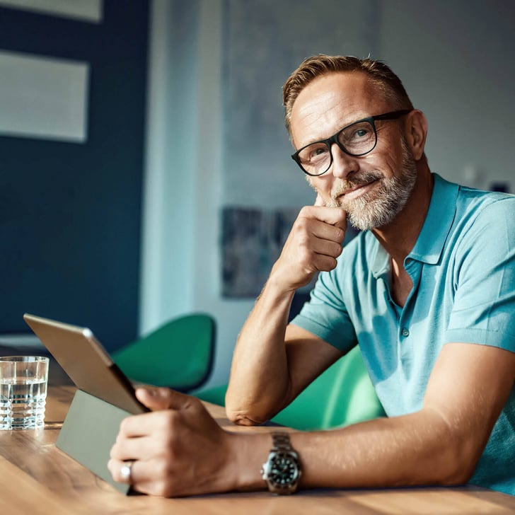 Mann mit Brille sitzt am Tisch und schaut nachdenklich auf ein Tablet.