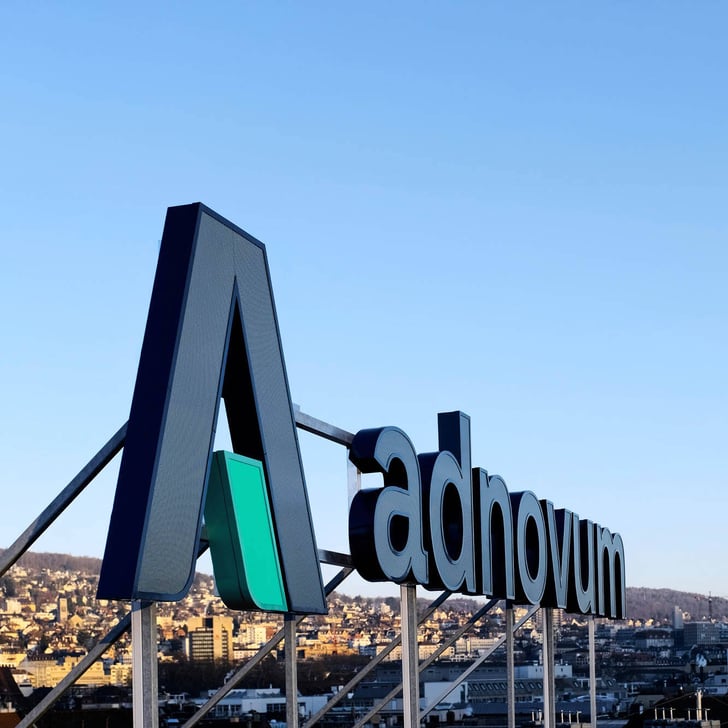Adnovum-Firmenschild auf einem Gebäude vor Stadtpanorama und blauem Himmel.