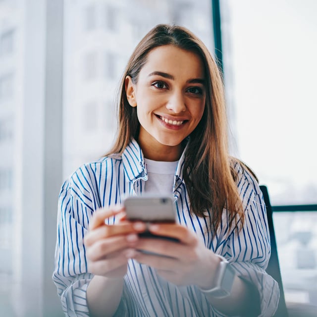Lächelnde Frau hält ein Smartphone in einem hellen Raum.