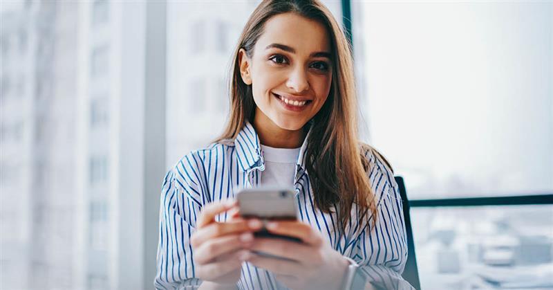 Lächelnde Frau hält ein Smartphone in einem hellen Raum.