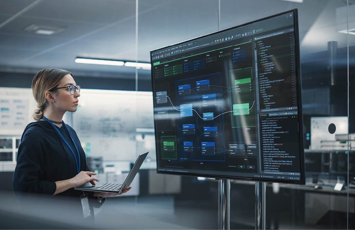 Eine Ingenieurin arbeitet an einem Laptop und überwacht gleichzeitig Anwendungs-Dashboards auf einem grossen Bildschirm in einem modernen Büro.